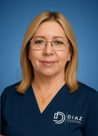 Una mujer con pelo rubio y gafas que lleva una camiseta azul marino con el logotipo de Diaz Dental, de pie delante de un fondo azul y sonriendo suavemente.